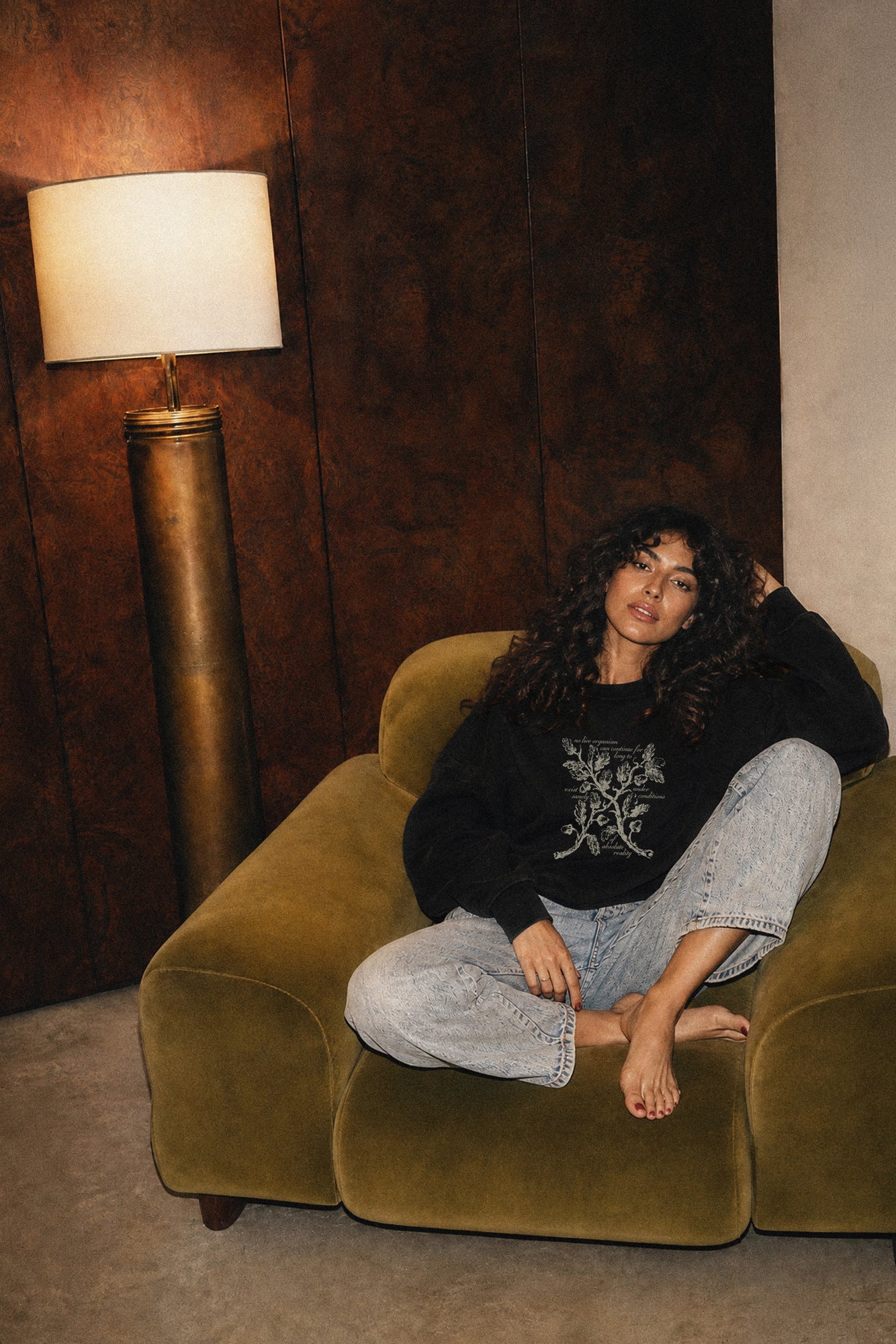 Woman sitting on a green armchair in a room with a lamp and dark wall, wearing a black crewneck sweatshirt with a floral design and Haunting of Hill House by Shirley Jackson quote