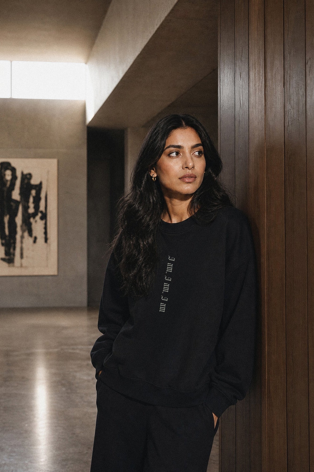 Woman wearing a black hoodie with text, standing in a modern interior setting.