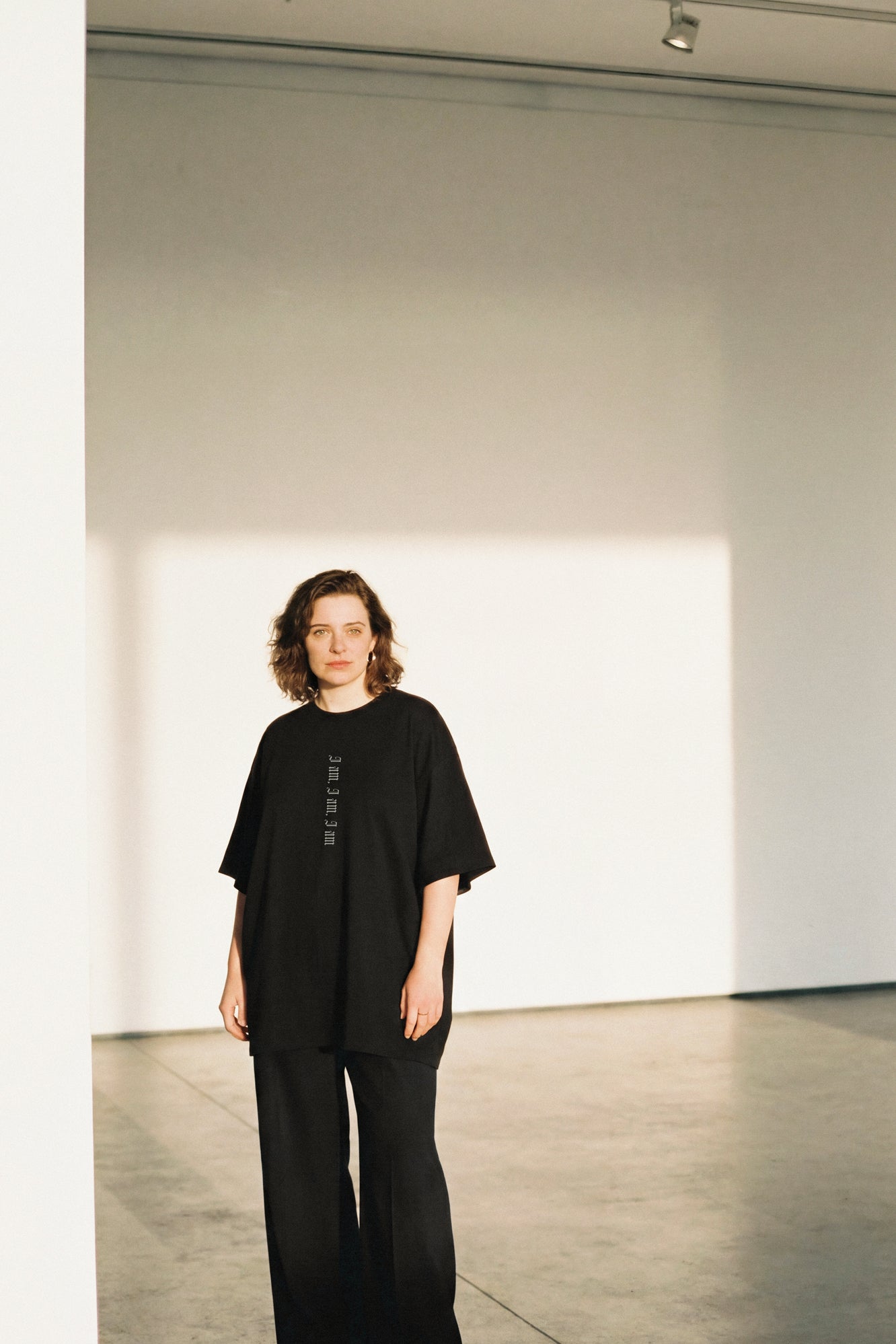 Person wearing a black outfit in a minimalistic room with white walls and gray floor.