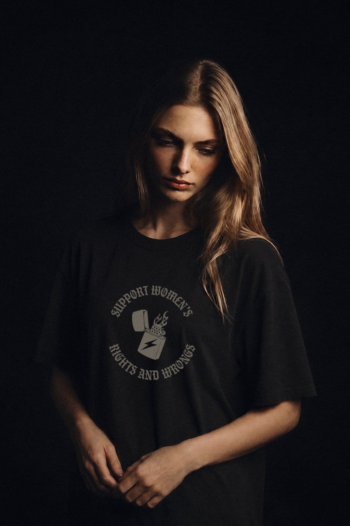 A woman with long light brown hair wearing a black graphic t-shirt with a circular white print that reads 'SUPPORT WOMEN'S RIGHTS AND WRONGS' around a small illustration, posed against a solid black background.