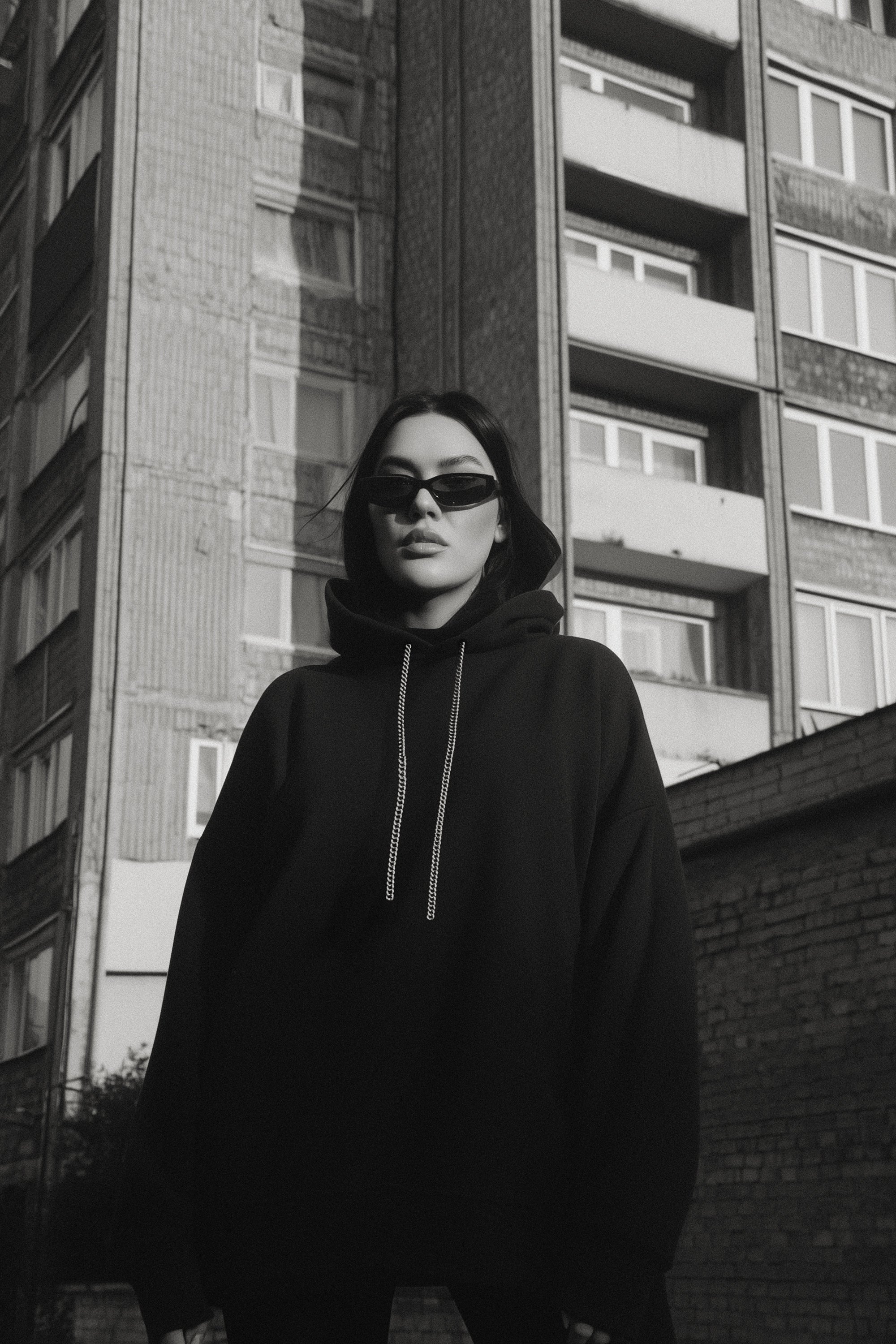 Person wearing a black hoodie and sunglasses standing in front of a tall building.