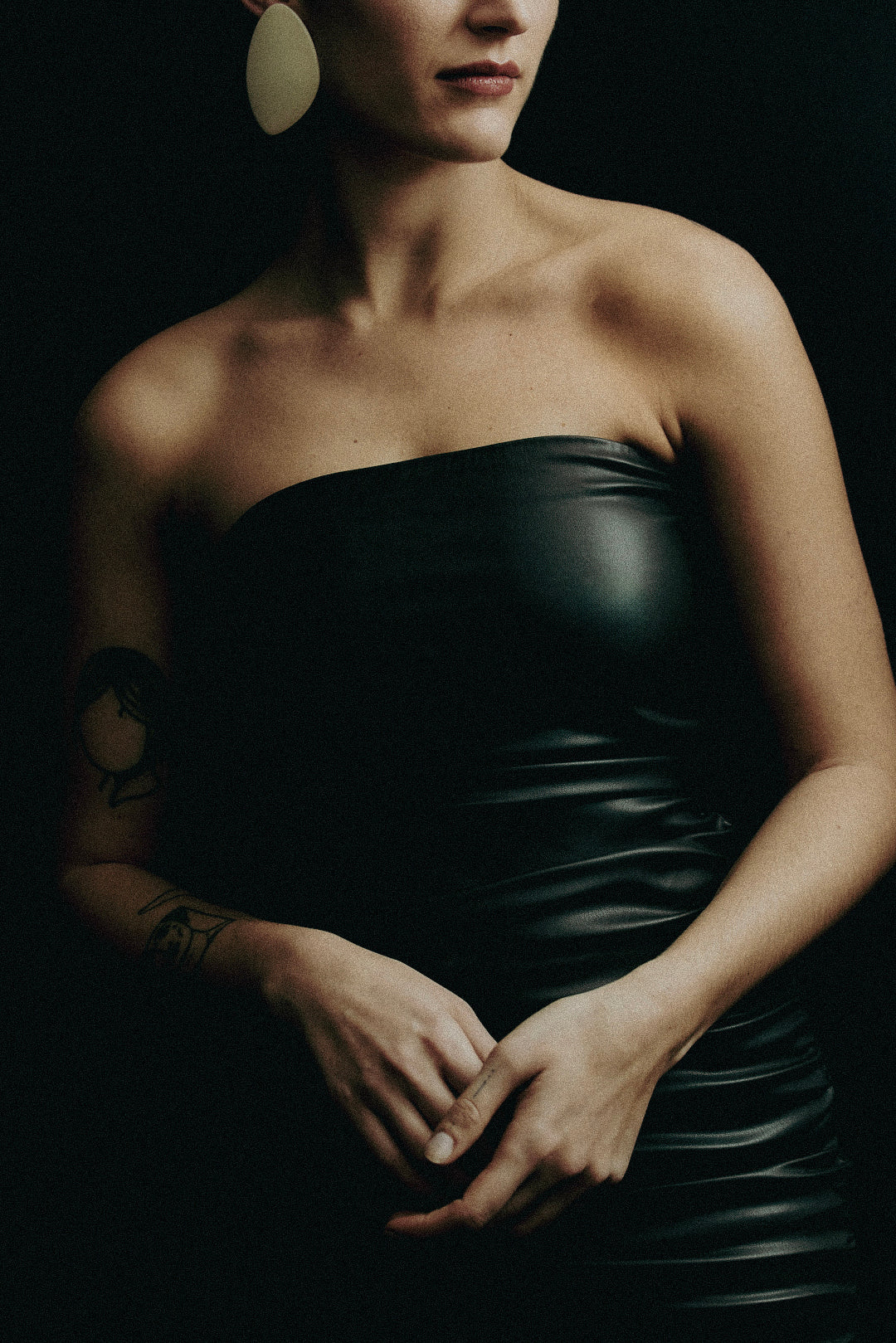A woman in her 30s wearing a black leather dress and edgy minimalist earrings standing against a moody dark background