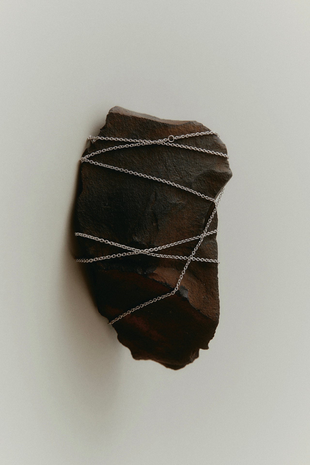 A dark, organic looking stone with a silver chain necklace wrapped around it against a light gray background