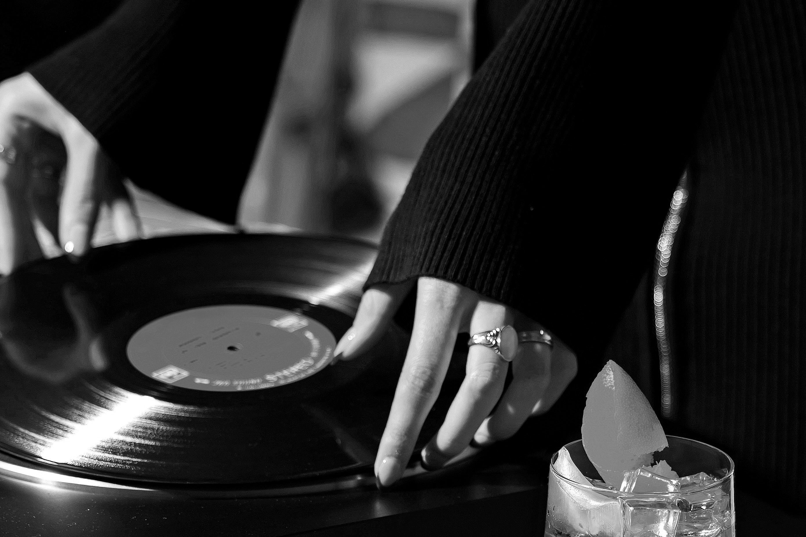 A closeup of a woman wearing a black sweater dress changing a vinyl record while drinking a cocktail