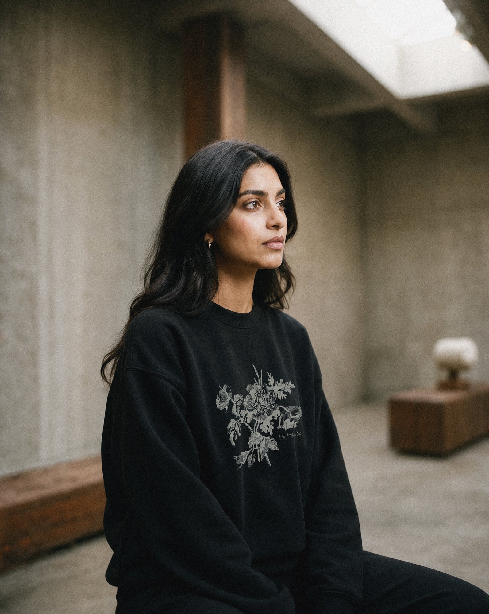 Woman wearing a black sweatshirt with floral design and the quote "et in arcadia ego" sitting in an indoor setting.
