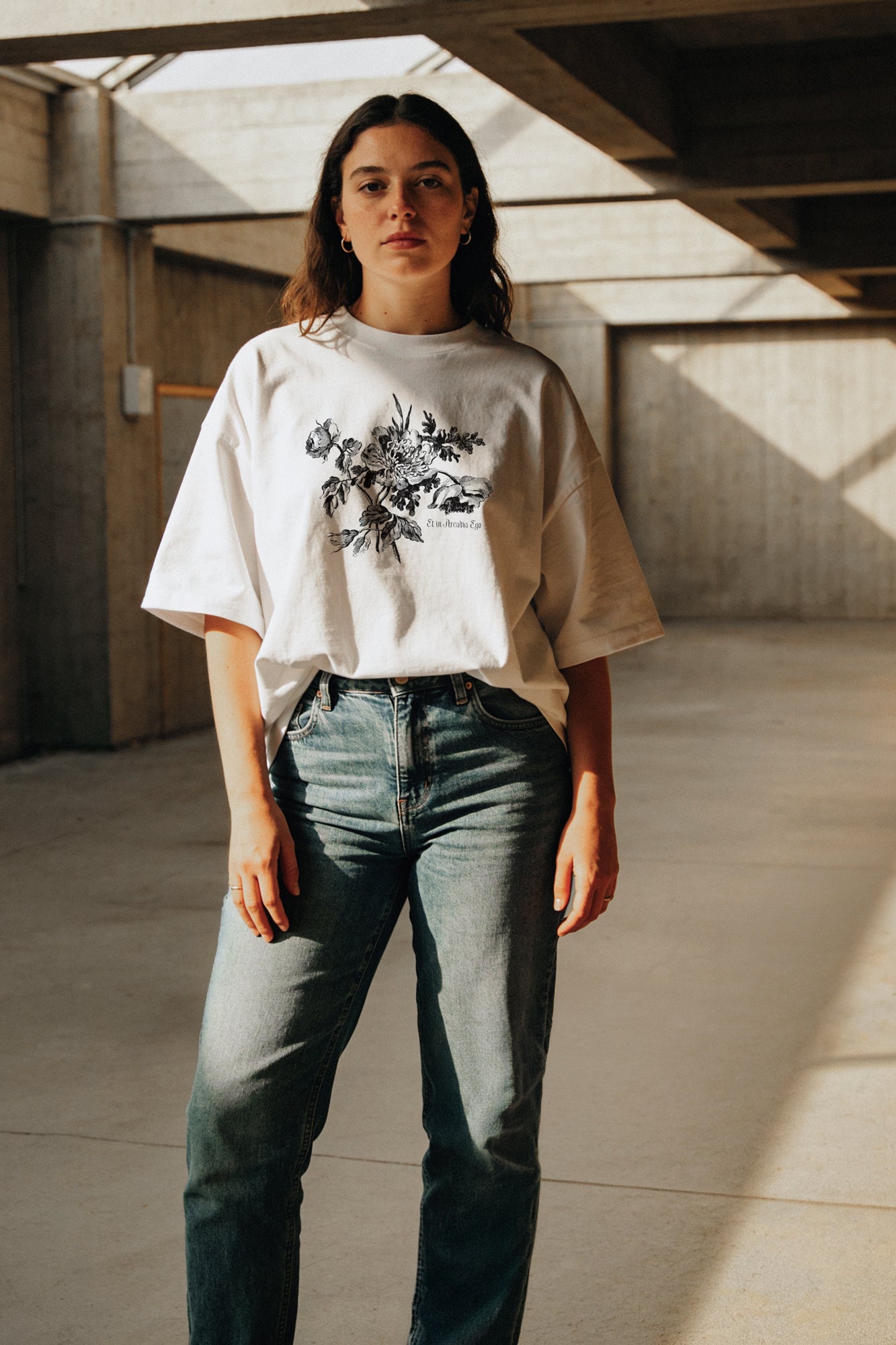 Person wearing a white t-shirt with a floral design and the quote "et in arcadia ego" and jeans in an indoor setting