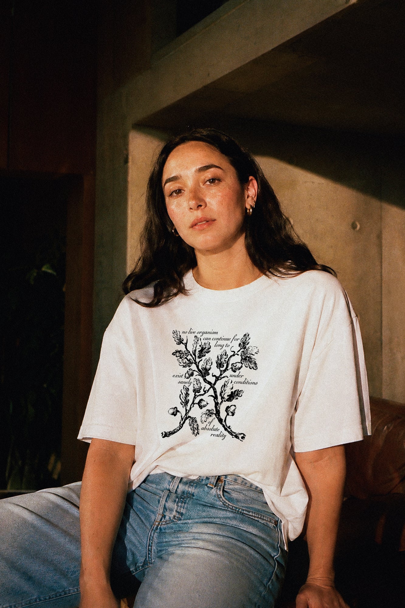 Person wearing a white t-shirt with black floral design and and a Haunting of Hill House by Shirley Jackson quote, sitting indoors.