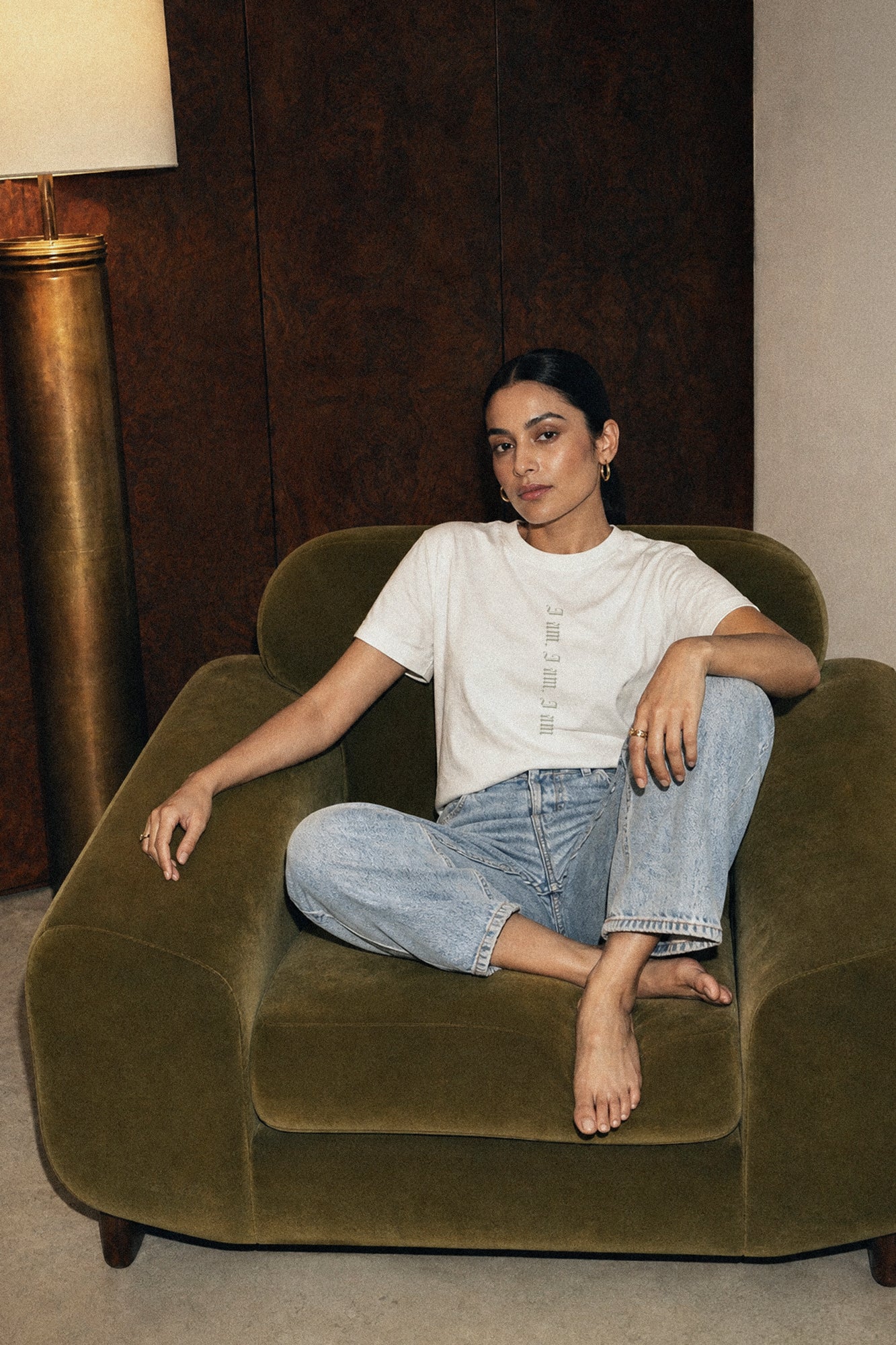 Woman sitting on a brown armchair in a room with a lamp and wall wearing a Sylvia Plath t-shirt.