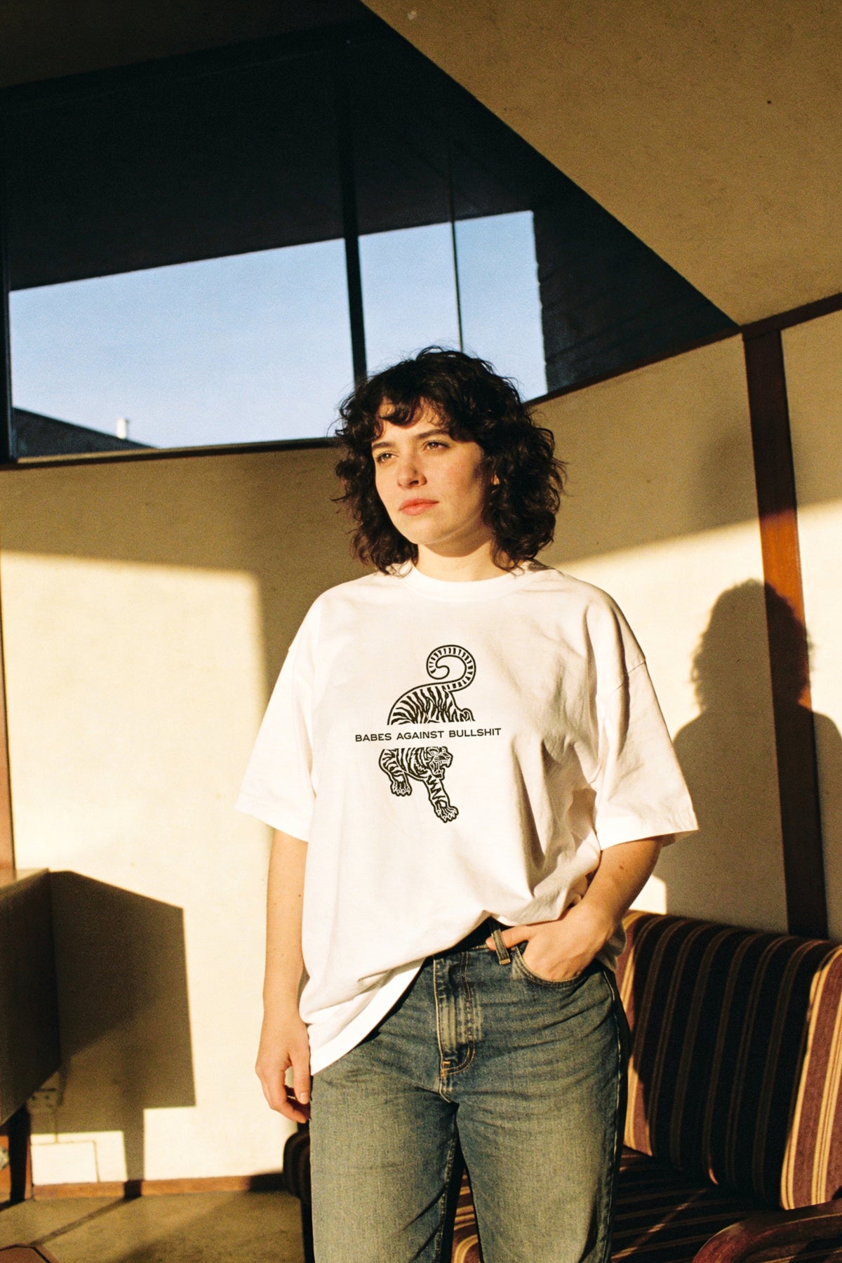 A person with curly dark hair stands indoors wearing an oversized white graphic T-shirt and blue jeans, with warm sunlight casting shadows on the wall behind them.