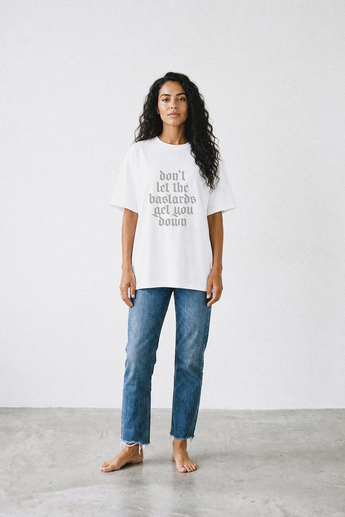 A woman stands barefoot in a white oversized graphic T-shirt and blue jeans against a plain light gray wall and concrete floor. The shirt has gray text reading, "don't let the bastards get you down.