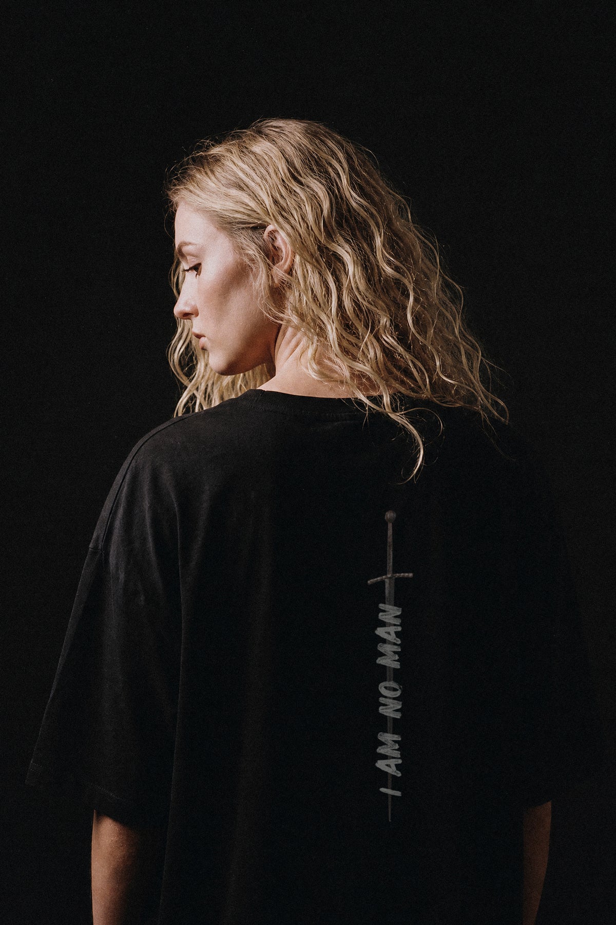 A woman with wavy blonde hair is shown from the back wearing a black oversized T-shirt with a vertical white graphic and the text 'I AM NO MAN' printed down the back, set against a solid black background.
