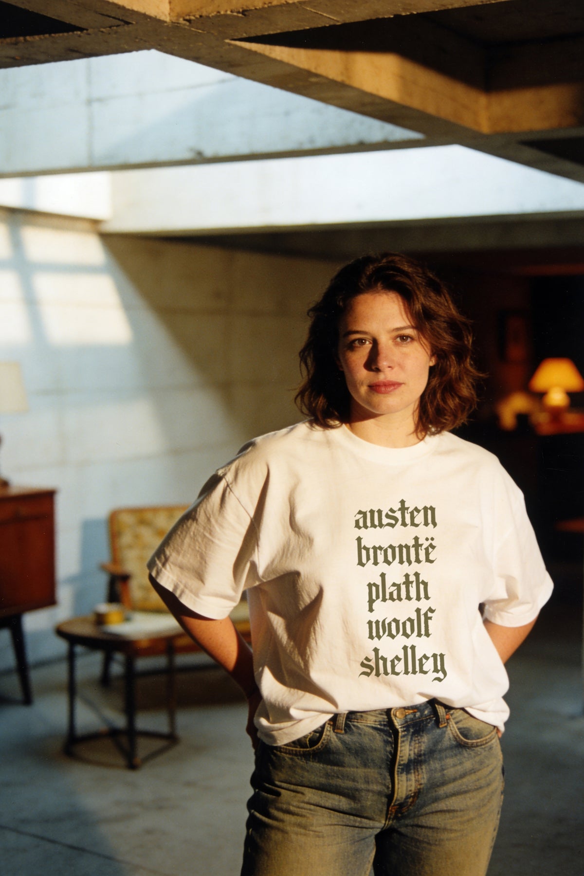 A woman with shoulder-length brown hair stands indoors wearing an oversized white graphic T-shirt with the names of Austen, Brontë, Plath, Woolf, and Shelley printed vertically in black serif text, paired with blue jeans.