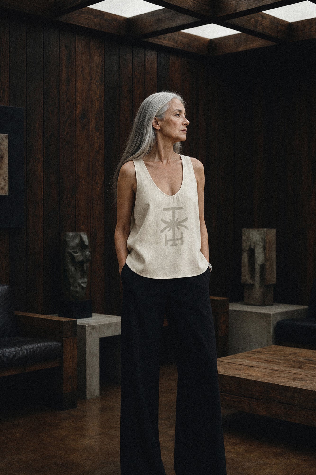 Woman standing in a dimly lit room with wooden walls and sculptures. Original artwork and clothing by Sara Kraus, Double Kick, alternative, small-batch, made-to-order, linen, artistic apparel and designs. Hand painted, custom tank top.
