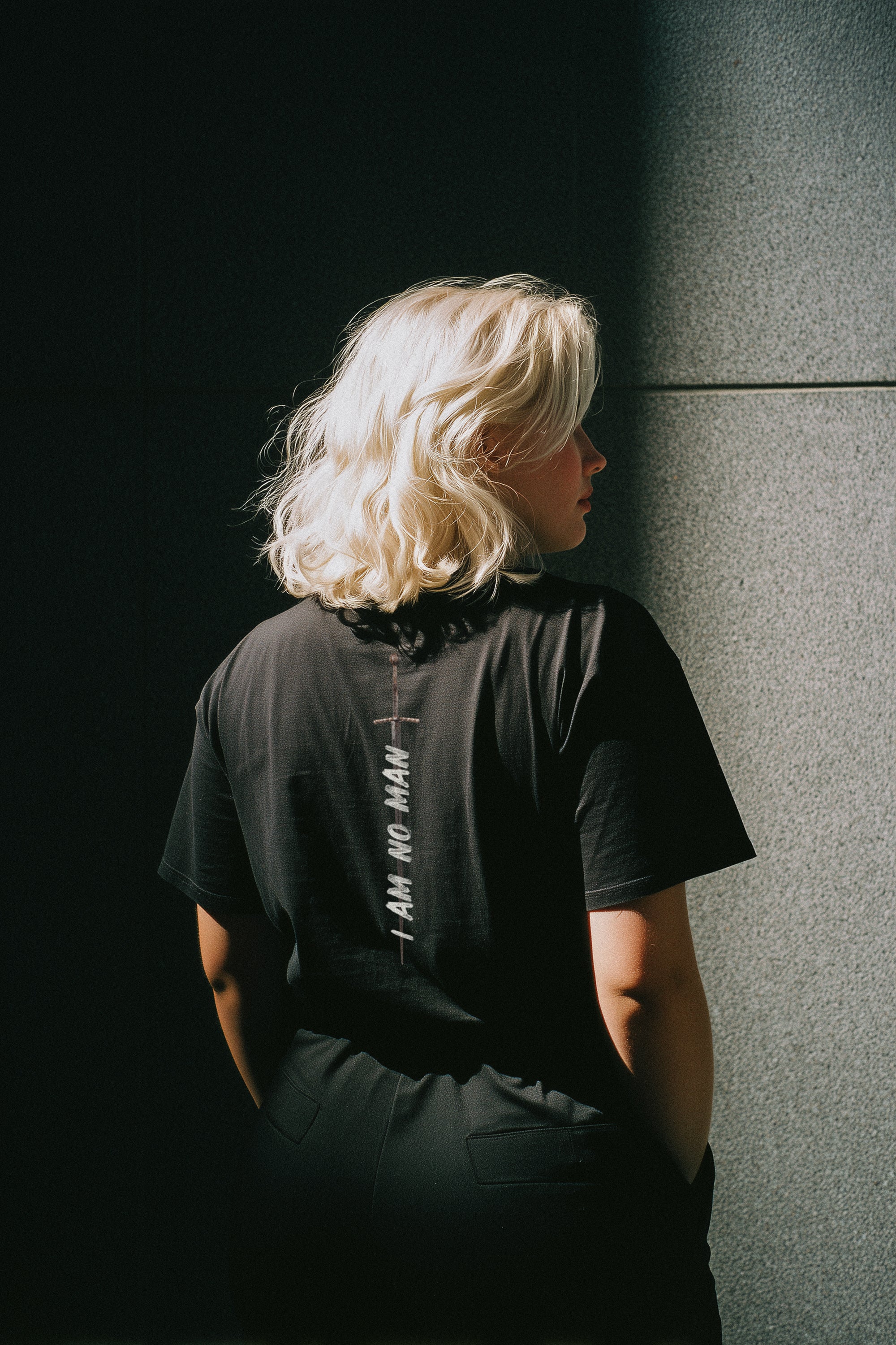 Person wearing a black t-shirt with text on the back, standing against a dark wall.