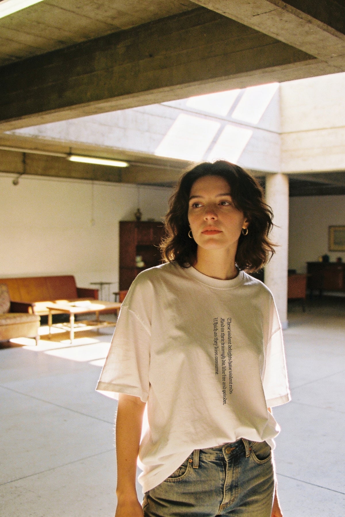 A woman wearing a white graphic t-shirt and blue jeans standing in a modern, sunlit concrete room.